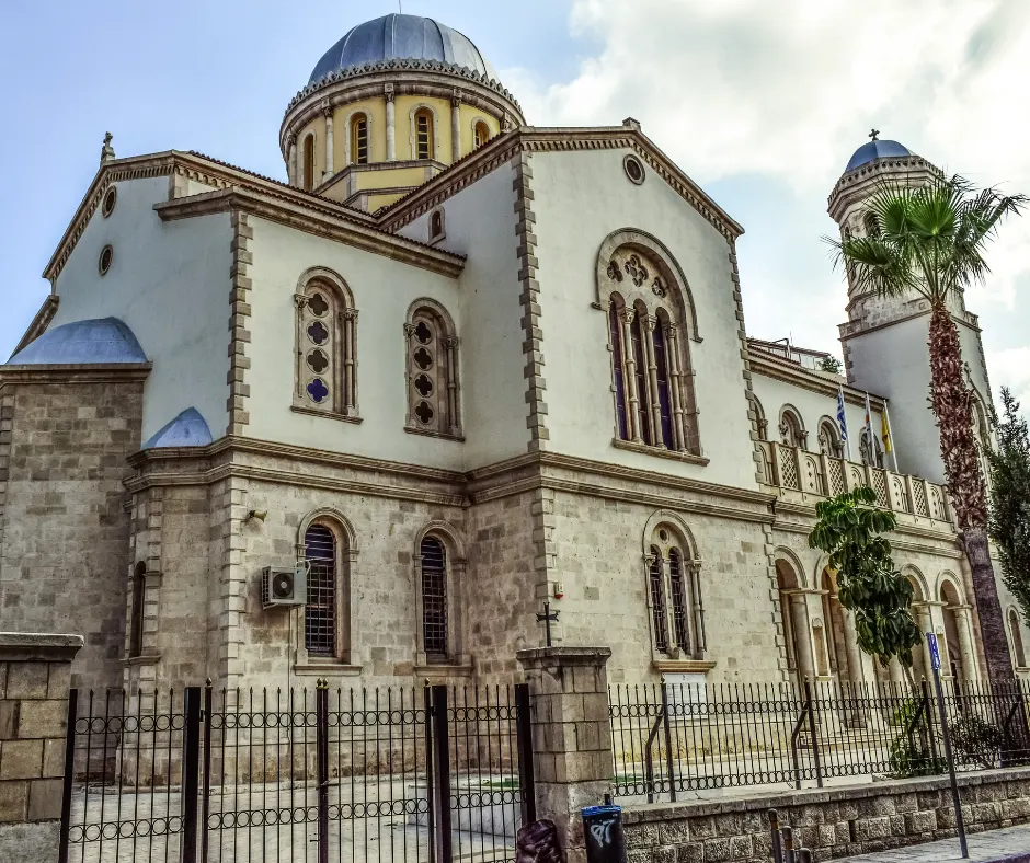 Ayia Napa Cathedral seen from the street — sandstone lower walls, rendered upper walls, arched and rose windows, a blue-grey dome and the bell tower beyond