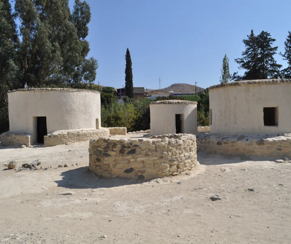 Three reconstructed round houses spaced across open ground, with a circular drystone wall base in the foreground