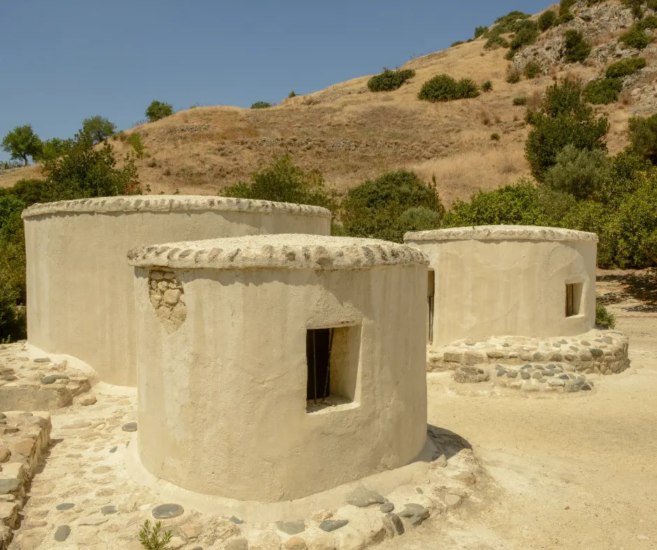 Three rebuilt round houses with domed stone-edged roofs, standing on pale ground below a dry scrub-covered hillside