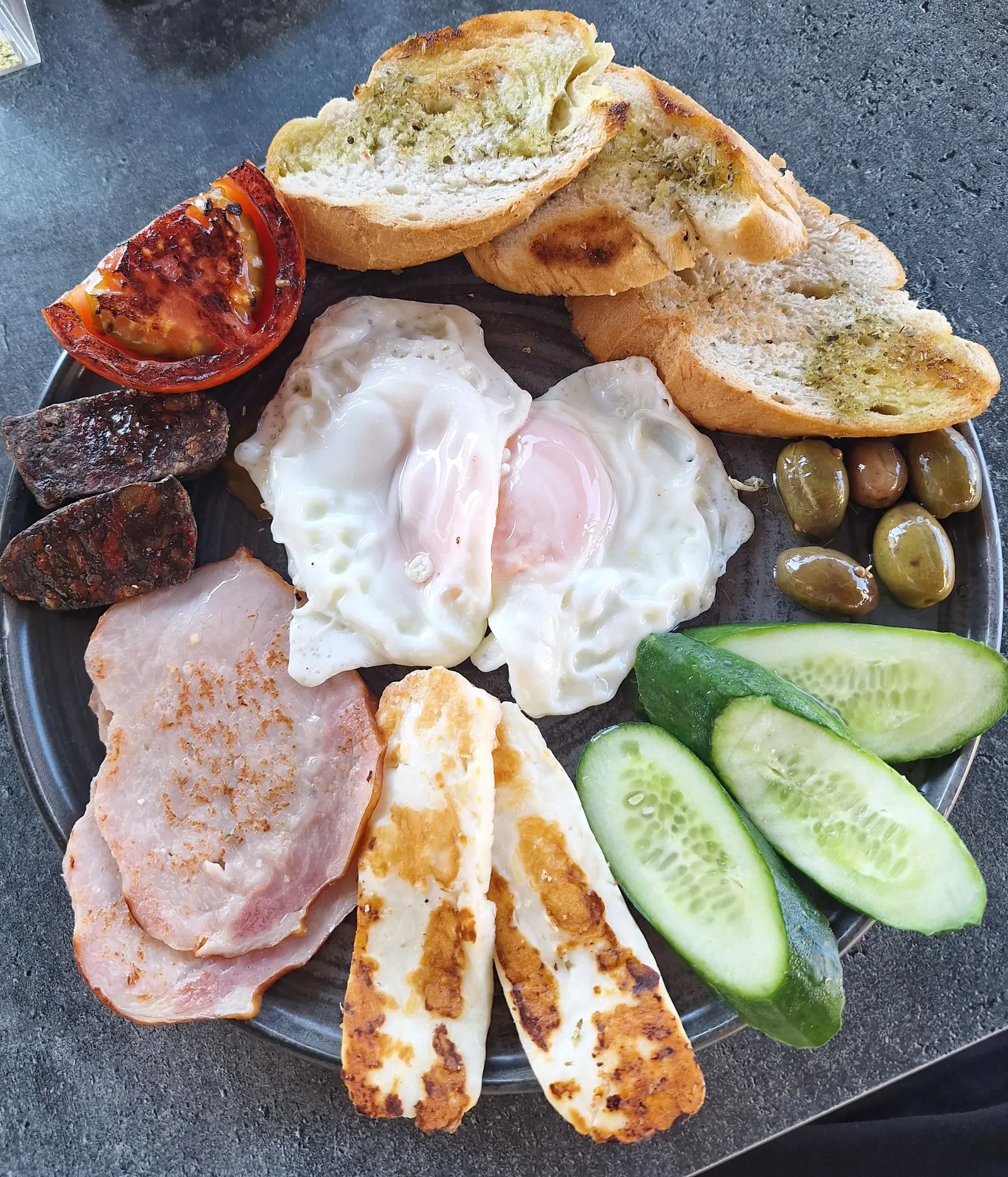 A Cypriot breakfast plate with fried eggs, grilled halloumi, ham, spicy sausage, olives, cucumber, grilled tomato and herbed bread